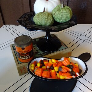 May include: A black pedestal cake stand with three white and green fabric pumpkins and a small can of curry powder on a book. A black and white bowl filled with orange and yellow candy corn sits on a black oval coaster.
