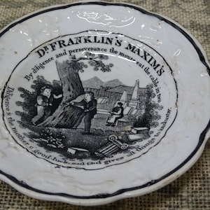 May include: A small, white ceramic plate with a black rim and a scalloped edge. The plate features a black and white illustration with text that reads "Dr. Franklin's Maxims". The illustration depicts figures near a tree and a building.