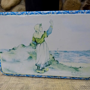 May include: Rectangular tin with a painted scene of a person waving near the sea. The person wears a traditional outfit with a white skirt and green top. The tin has a blue floral border.