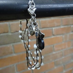 May include: Silver-toned bracelet with multiple charms, including a heart, a flower, and a black stone. The bracelet also features strands of white beads strung on brown cord. The background is a brick wall.