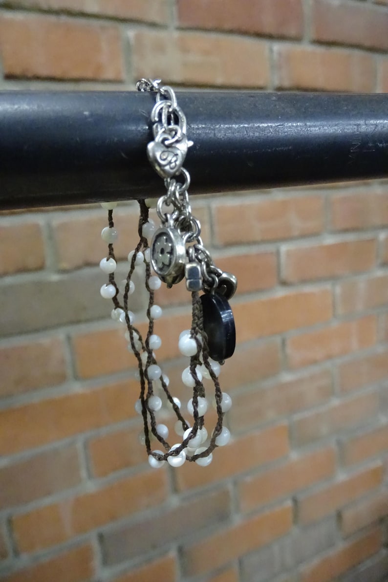 May include: Silver-toned bracelet with multiple charms, including a heart, a flower, and a black stone. The bracelet also features strands of white beads strung on brown cord. The background is a brick wall.