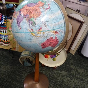 Może przedstawiać: Stary globus na drewnianym stojaku z podstawą w kolorze miedzi. Globus przedstawia szczegółową mapę z różnymi krajami w różnych kolorach. Stojak jest w kolorze jasnobrązowym. To klasyczny przedmiot edukacyjny.