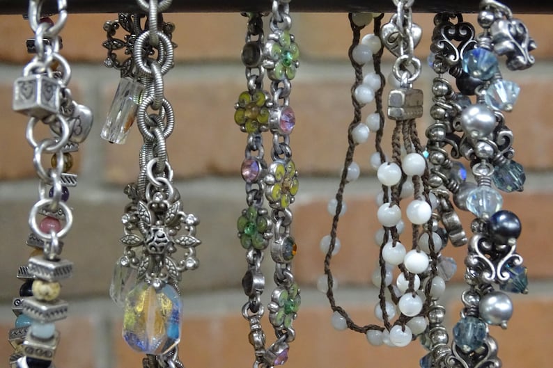 May include: A collection of silver-toned charm bracelets and necklaces with various decorative elements. The charms include beads, crystals, and floral designs. Some necklaces feature white beads and others have colorful accents.