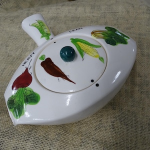 Puede incluir: Un recipiente de cerámica blanca con tapa, que presenta un pomo verde e imágenes pintadas de verduras. La tapa tiene las palabras "Heavy" y "Lean" impresas, con imágenes de remolacha, zanahoria, maíz y judías verdes.