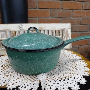 Może przedstawiać: Stary, zielony garnek emaliowany z pokrywką i czarnym uchwytem. Garnek stoi na białej koronkowej serwetce. Etykieta przymocowana do uchwytu głosi "Warmth & Cheer".