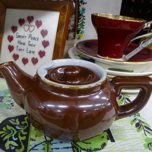 Puede incluir: Una tetera de cerámica marrón con borde y tapa blancos, colocada sobre un mantel estampado. Una taza de té roja con borde dorado se encuentra sobre una pila de platillos blancos. Un bordado enmarcado con el texto "Great Peace Have They That Love" está en el fondo.