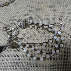 May include: A multi-strand bracelet featuring small, white beads strung on dark brown cord. The bracelet has a silver-toned clasp and decorative charms, including a flower and a black oval bead. The jewelry rests on a textured, tan surface.