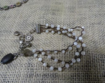 Könnte beinhalten: Ein mehrreihiges Armband mit kleinen, weißen Perlen auf dunkelbraunem Kordelband. Das Armband hat einen silberfarbenen Verschluss und dekorative Anhänger, darunter eine Blume und eine schwarze ovale Perle. Der Schmuck liegt auf einer strukturierten, beigefarbenen Oberfläche.
