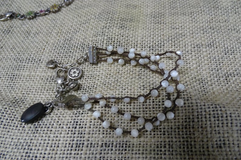 May include: A multi-strand bracelet featuring small, white beads strung on dark brown cord. The bracelet has a silver-toned clasp and decorative charms, including a flower and a black oval bead. The jewelry rests on a textured, tan surface.