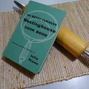 May include: A green cookbook titled "The Betty Furness Westinghouse Cook Book" with a white illustration of a frying pan. The book is on a woven placemat with a wooden rolling pin.