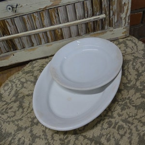 Peut inclure: Deux assiettes blanches, ovales et rondes, empilées l'une sur l'autre. Les assiettes présentent une légère décoloration sur les bords. Les assiettes sont posées sur un tissu à motifs.