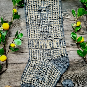 Può includere: Un calzino lavorato a maglia con un motivo geometrico in tonalità di grigio e beige. Il calzino è appoggiato su una superficie di legno con una piccola ciotola e gomitoli di lana nelle vicinanze.