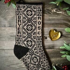 Puede incluir: Una media de Navidad de punto negro y beige con los nombres "BRYER" y "ROSE" bordados en la parte delantera. La media está sobre una superficie de madera con follaje verde y bayas rojas alrededor.