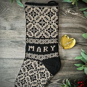 May include: A black and beige knitted Christmas stocking with the name "MARY" embroidered on the front. The stocking is on a wooden background with green foliage and red berries.