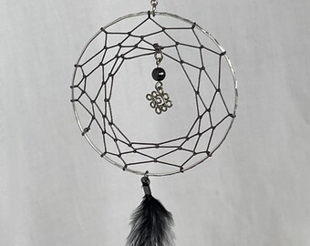 May include: A handcrafted dreamcatcher with a silver-colored metal frame and a web of black string. A small, decorative charm hangs in the center, and a black and gray feather dangles from the bottom. The dreamcatcher is suspended by a silver chain.