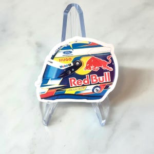 May include: A sticker featuring a racing helmet design with the Red Bull logo. The helmet has blue, red, yellow, and white accents, with the text "Red Bull" prominently displayed. Additional text includes "Ford Racing", "Hugo", and "Mobil 1".