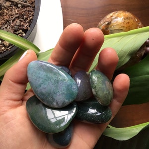 May include: A collection of six green, blue, and grey agate stones, tumbled and polished smooth. The stones are arranged in a hand, with the largest stone in the center.
