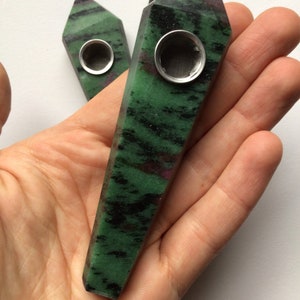 May include: A green and black crystal pipe with a metal bowl and screen. The pipe is shaped like a long, thin point.
