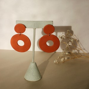 Puede incluir: Un par de pendientes de color terracota exhibidos en un soporte gris claro. Cada pendiente presenta un pequeño perno redondo conectado a un colgante circular más grande con un agujero en el centro. Diseño minimalista y moderno.