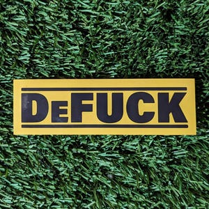 May include: A yellow rectangular sticker with black text that reads "DeFUCK". The sticker has a black border and is set against a background of green artificial turf.