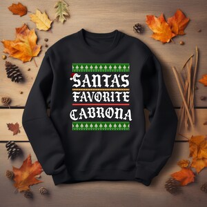 May include: Black sweatshirt with the text "SANTA'S FAVORITE CABRONA" in white, red, and gold. The design includes green and white Christmas tree patterns at the top and bottom, with a Santa hat on the letter "S".