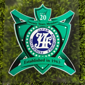 May include: A green and black logo with a crown and the text "Twenty Years Member" and "Established in 1963". The logo also features a blue circle with the text "Japan Automobile Federation" and the letters "JAF" in a stylized font.