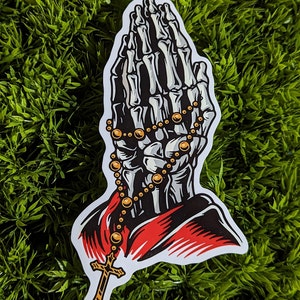 May include: A sticker of a skeleton hand in a praying position with a gold rosary and cross.