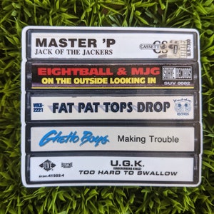 May include: A sticker featuring a stack of five cassette tapes with various album titles and artist names. The titles include "Master P," "Eightball & MJG," "Fat Pat," "Ghetto Boys," and "U.G.K." The background is a green, grassy texture.