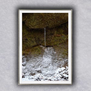 以下が含まれることがあります： 自然の風景を写した額縁写真。小さな水の流れが、苔で覆われた岩壁を流れ落ちています。画像の下部は雪で覆われており、緑、茶色、白、透明な水のコントラストを生み出しています。