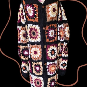 May include: A long, button-down crochet cardigan in a patchwork design. The jacket features a black collar, cuffs, and borders, with square panels in shades of white, burgundy, orange, and tan. The cardigan has wooden buttons and a floral pattern on the mannequin.