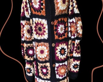 May include: A long, button-down crochet cardigan in a patchwork design. The jacket features a black collar, cuffs, and borders, with square panels in shades of white, burgundy, orange, and tan. The cardigan has wooden buttons and a floral pattern on the mannequin.