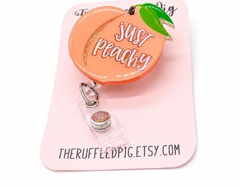 May include: A peach-shaped badge reel featuring a light orange colour, a green leaf, and the text "just peachy" in white script. The badge reel is attached to a clear plastic strap with a small, round, glittery button, mounted on a light pink card.