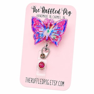 May include: A butterfly-shaped badge reel featuring a pink, blue, and white glittery resin design. The badge reel is attached to a clear plastic strap with a small circular pink glitter accent. The display card reads The Ruffled Pig.