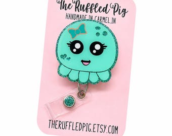 Può includere: Un portabadge a forma di medusa verde menta glitterata con un fiocco color ottanio, grandi occhi neri e una bocca sorridente. Il badge è fissato a un cinturino in plastica trasparente con un bottone glitterato. La carta mostra il testo The Ruffled Pig.