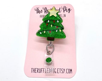 May include: A green glitter Christmas tree with white and red lights and a gold star on top. The tree is attached to a clear retractable badge holder with a green glitter circle on the bottom. The card reads "The Ruffled Pig Handmade" and "theruffledpig.etsy.com".