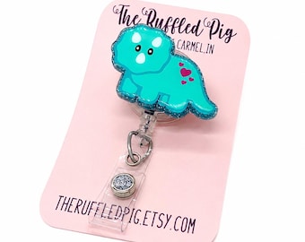Può includere: Un porta badge a forma di triceratopo turchese con dettagli glitterati e piccoli cuori rosa sul corpo. Il porta badge è fissato a un cinturino in plastica trasparente e a una clip in metallo argentato, su una carta rosa The Ruffled Pig.