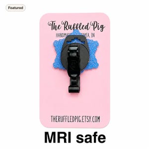 May include: A blue glittery badge reel with a black plastic clip attached to a pink display card. The card features the text The Ruffled Pig HANDMADE IN MUNCIE, IN and THERUFFLEDPIG.ETSY.COM. The badge reel is labeled as MRI safe.