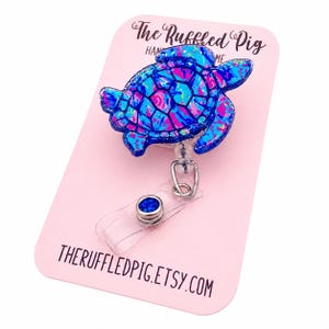 May include: A blue, pink, and purple sea turtle badge reel on a pink card. The turtle has a glittery finish. The card has the text "The Ruffled Pig" and "THERUFFLEDPIG.ETSY.COM". The reel has a clear plastic strap and a blue gem.