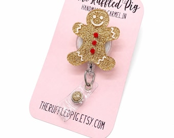 May include: A gold glitter gingerbread man badge reel attached to a light pink display card. The card features the text The Ruffled Pig, Handmade in Carmel, IN, and THERUFFLEDPIG.ETSY.COM. The gingerbread man has white icing details and three red buttons.