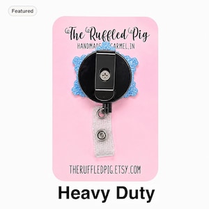 May include: A blue glittery badge reel with a black circular housing and a metal belt clip. The reel is attached to a clear plastic strap with a silver snap button. The product is mounted on a pink display card labeled The Ruffled Pig.