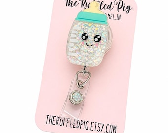 May include: A baby bottle-shaped badge reel with a smiling face, filled with glitter. The reel has a light blue lid and a yellow nipple. The badge reel is attached to a clear plastic holder and a silver clip. The background is a pink card with text.
