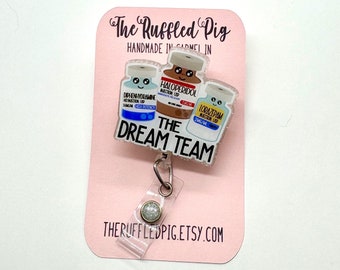 Op de afbeelding: Een roze kaart met de tekst "The Ruffled Pig" en "HANDMADE IN GARMEL IN" in zwart. De kaart heeft een transparante badgehouder met een glinsterende charme en een transparante charme met drie cartoon-medicijnflessen met gezichten en de tekst "THE DREAM TEAM".