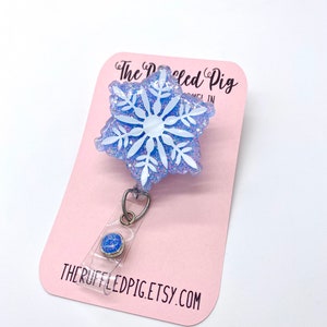 May include: A badge reel featuring a light blue, glittery, snowflake-shaped topper with a white snowflake design. The badge reel is attached to a pink display card printed with the text The Ruffled Pig and THERUFFLEDPIG.ETSY.COM.