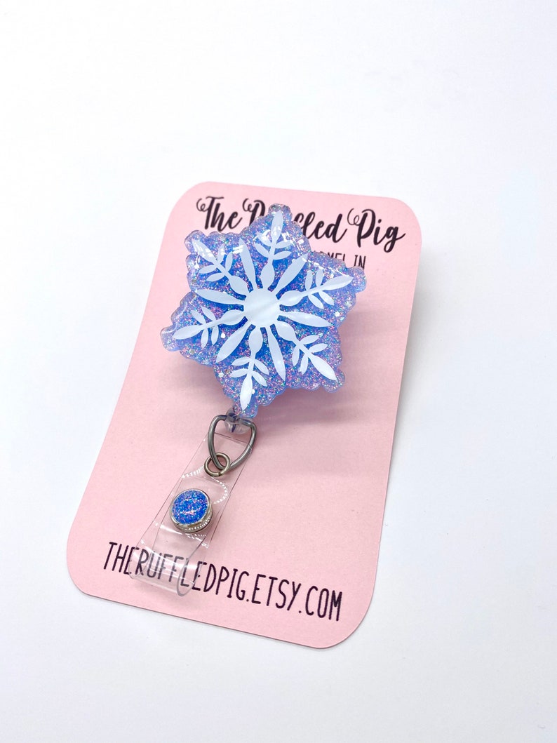 May include: A badge reel featuring a light blue, glittery, snowflake-shaped topper with a white snowflake design. The badge reel is attached to a pink display card printed with the text The Ruffled Pig and THERUFFLEDPIG.ETSY.COM.