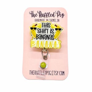 May include: A yellow and white badge reel with the text "THIS SHIFT IS BANANAS" and two cartoon bananas wearing sunglasses. The badge reel is attached to a pink card with the text "The Ruffled Pig" and "HANDMADE IN CARMEL, IN".