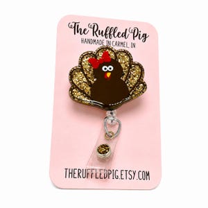 May include: A turkey badge reel featuring a brown turkey with a red bow, white eyes, and a yellow beak, set against a gold glitter fan-shaped background. The badge is attached to a clear plastic strap and a circular gold glitter button on a pink card.