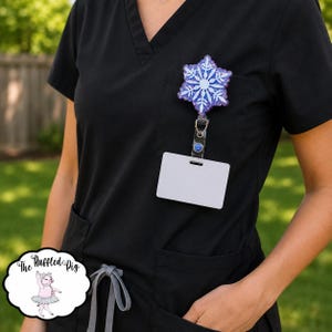 May include: A badge reel featuring a glittery purple and blue snowflake design attached to a black scrub top. The snowflake has a white center and white crystalline branches. A blank white rectangular ID card holder hangs from the metal clip.