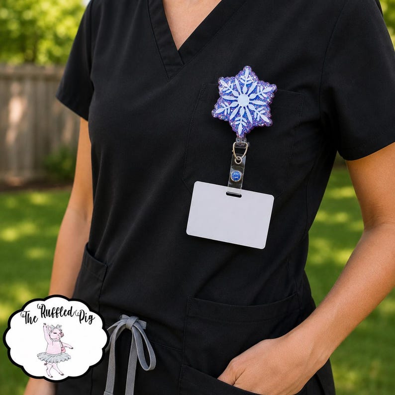 May include: A badge reel featuring a glittery purple and blue snowflake design attached to a black scrub top. The snowflake has a white center and white crystalline branches. A blank white rectangular ID card holder hangs from the metal clip.