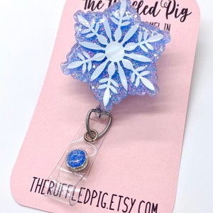 May include: A badge reel featuring a blue snowflake shape with white snowflake detailing and iridescent glitter embedded in the resin. The badge reel has a clear plastic strap with a small blue glitter button and is attached to a pink display card.
