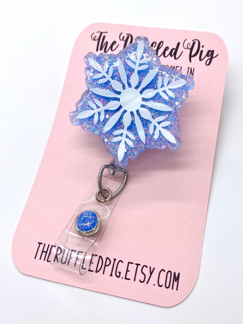 May include: A badge reel featuring a blue snowflake shape with white snowflake detailing and iridescent glitter embedded in the resin. The badge reel has a clear plastic strap with a small blue glitter button and is attached to a pink display card.